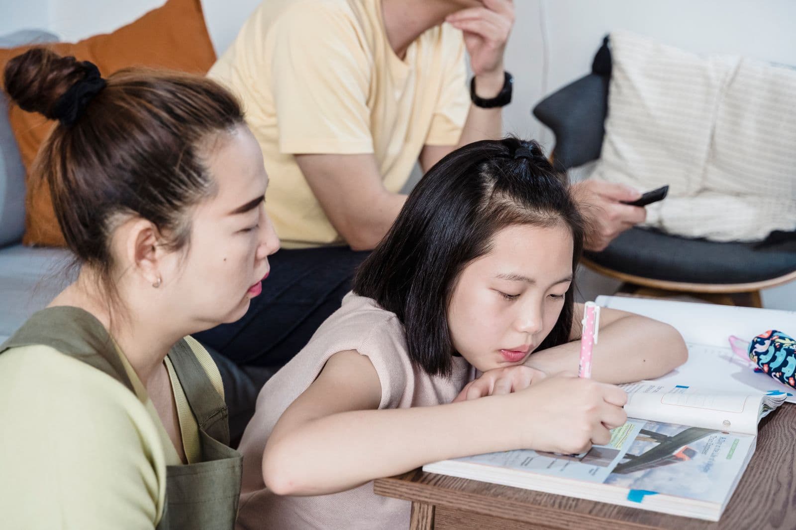 A girl slumped over her homework at the table while her mom sits beside her and her dad is distracted on his phone in the background